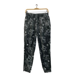 Può includere: Pantaloni della tuta in velluto grigio scuro con vita con coulisse, con un motivo ripetuto di illustrazioni di gatti dei cartoni animati bianchi e stelle. I pantaloni sono appesi a una gruccia di legno.