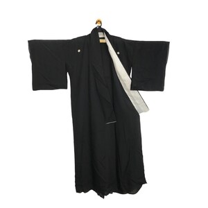 May include: A black kimono with wide sleeves hangs on a wooden hanger. The kimono has a white lining visible at the front and two white buttons. The garment is made of a smooth, dark fabric.