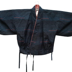 Könnte beinhalten: Eine dunkel marineblaue kurze Jacke mit weiten Ärmeln und einem dezenten horizontalen Muster aus dünnen, rostfarbenen Linien. Das Kleidungsstück hat einen V-Ausschnitt mit rostfarbenem Innenfutter und zwei dünne Stoffbänder am unteren Saum.