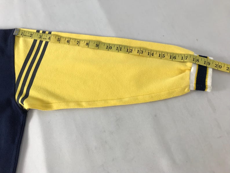 May include: A long-sleeved shirt with a navy blue body and a yellow sleeve. The yellow sleeve has three black stripes and a white and navy blue striped cuff. A measuring tape shows the sleeve is approximately 20 inches long.