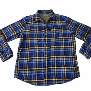 Puede incluir: Una camisa de franela a cuadros azules, amarillos y negros con botones y mangas largas. La camisa tiene dos bolsillos en el pecho y un forro gris suave. La camisa está tendida sobre un fondo blanco.