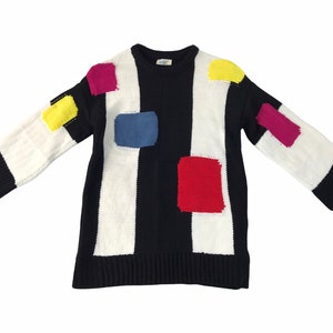 Puede incluir: Un suéter de manga larga con base negra, rayas blancas y parches coloridos. El suéter presenta parches rectangulares amarillos, azules, rojos y magenta. Las mangas y el dobladillo son negros.