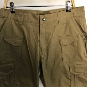Pu&ograve; includere: Pantaloni cargo color kaki con pi&ugrave; tasche, tra cui due tasche laterali e due tasche frontali. I pantaloni sono appesi a una gruccia di legno su sfondo bianco. I pantaloni hanno una chiusura con bottoni e una cerniera sulla tasca laterale.
