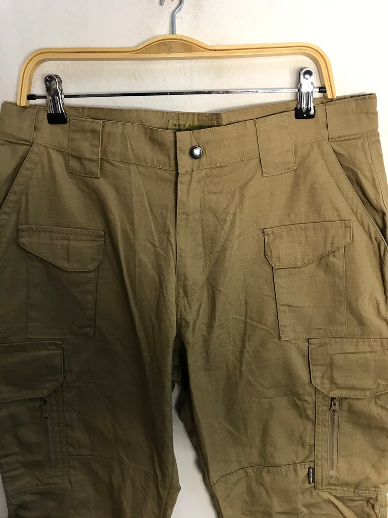 Pu&ograve; includere: Pantaloni cargo color kaki con pi&ugrave; tasche, tra cui due tasche laterali e due tasche frontali. I pantaloni sono appesi a una gruccia di legno su sfondo bianco. I pantaloni hanno una chiusura con bottoni e una cerniera sulla tasca laterale.