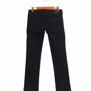 Könnte beinhalten: Schwarze Jeans aus Denim mit klassischem Fünf-Taschen-Design, mit Gesäßtaschen mit Knopfverschluss und einem Leder-Markenaufnäher. Die Jeans hängen an einem Holzbügel vor weißem Hintergrund.
