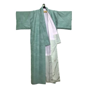 Può includere: Un kimono verde chiaro con un motivo floreale, appeso a una gruccia di legno. Il kimono è aperto, rivelando una fodera bianca. Le maniche sono ampie e il tessuto ha una leggera lucentezza.