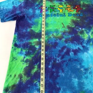 Puede incluir: Una camiseta tie-dye con un esquema de color azul, verde y morado. La camiseta presenta un gr&aacute;fico de cinco osos bailando con el texto "Grateful Dead" debajo. La camiseta se mide con una cinta m&eacute;trica, mostrando una longitud de 74 cm.
