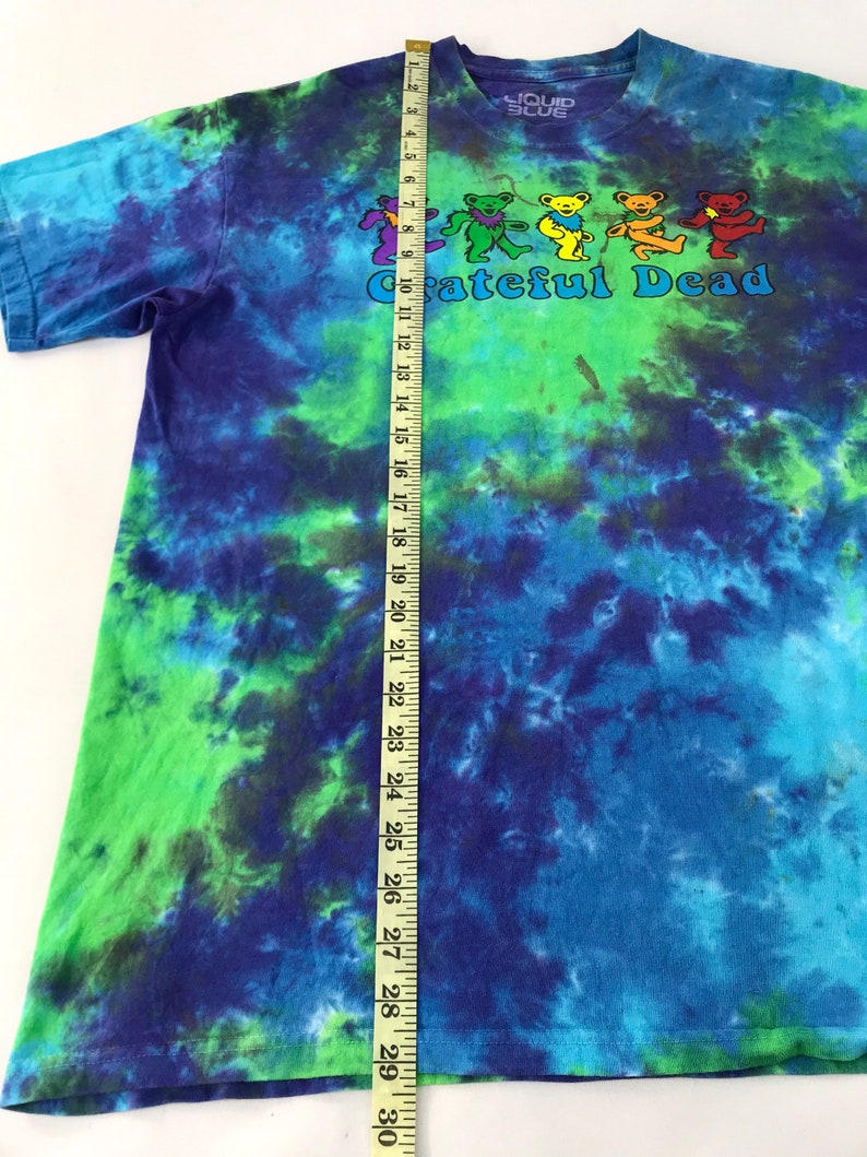 Puede incluir: Una camiseta tie-dye con un esquema de color azul, verde y morado. La camiseta presenta un gr&aacute;fico de cinco osos bailando con el texto "Grateful Dead" debajo. La camiseta se mide con una cinta m&eacute;trica, mostrando una longitud de 74 cm.