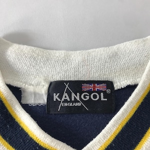 May include: Close-up of a navy blue shirt with a white collar and yellow stripes. The shirt features a black tag with the brand name "KANGOL ENGLAND" and the Union Jack flag.