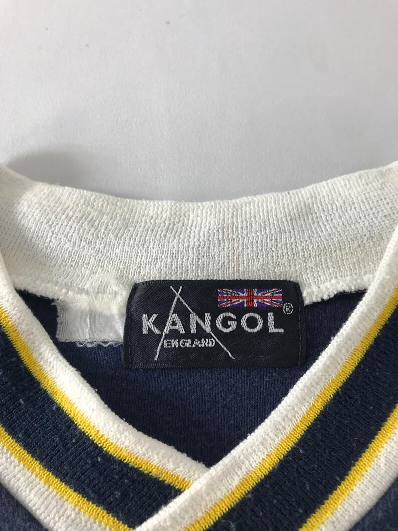 May include: Close-up of a navy blue shirt with a white collar and yellow stripes. The shirt features a black tag with the brand name "KANGOL ENGLAND" and the Union Jack flag.