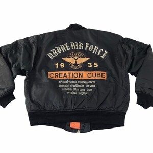 May include: Black bomber jacket with embroidered "NAVAL AIR FORCE" text and a winged emblem. The back of the jacket features the text "CREATION CUBE" and additional text. The jacket has a black ribbed collar and cuffs.