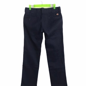Könnte beinhalten: Marineblaue Arbeitshose, die an einem grünen Kleiderbügel hängt. Die Hose hat ein klassisches, gerades Bein-Design mit einem kleinen Logo-Patch auf der Gesäßtasche. Der Stoff scheint ein strapazierfähiges Arbeitsmaterial zu sein.