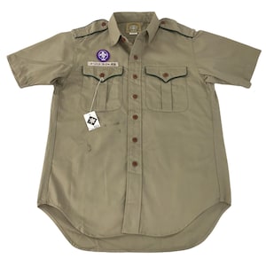 May include: Tan short-sleeved shirt with two chest pockets and a button-down front. The shirt has a collar and epaulets. A patch with a purple emblem and Japanese text is on the left pocket. A tag with a logo hangs from the pocket.