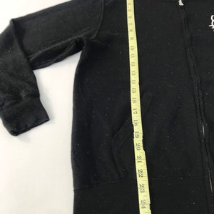 K&ouml;nnte beinhalten: Schwarzer Kapuzenpullover mit Rei&szlig;verschluss, Kapuze und Vordertaschen. Der Hoodie wird mit einem gelben Ma&szlig;band gemessen. Die Messung ist ca. 66 cm lang. Der Hoodie hat ein wei&szlig;es, gesprenkeltes Muster.
