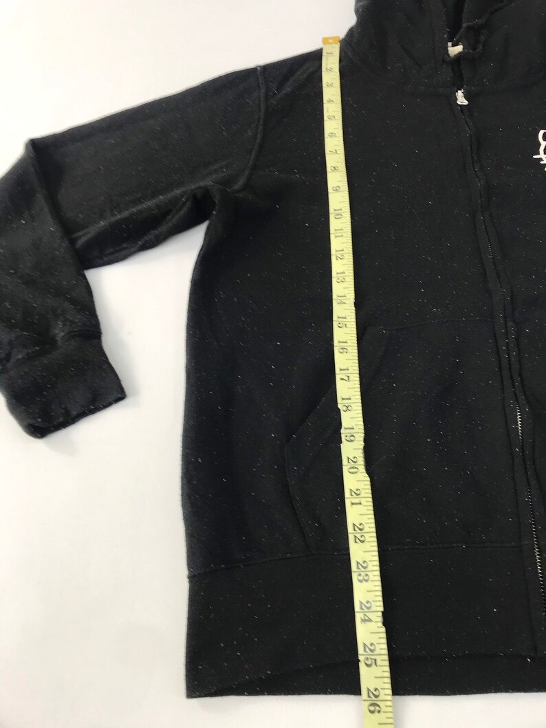 K&ouml;nnte beinhalten: Schwarzer Kapuzenpullover mit Rei&szlig;verschluss, Kapuze und Vordertaschen. Der Hoodie wird mit einem gelben Ma&szlig;band gemessen. Die Messung ist ca. 66 cm lang. Der Hoodie hat ein wei&szlig;es, gesprenkeltes Muster.