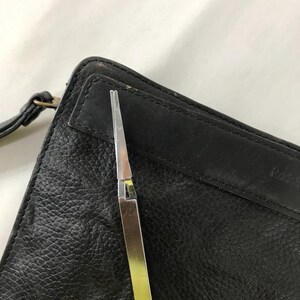 May include: A silver and gold metal tweezer tool is positioned over a black leather wallet. The wallet has a textured surface and a strap. The tweezer is open, revealing its pointed tips.