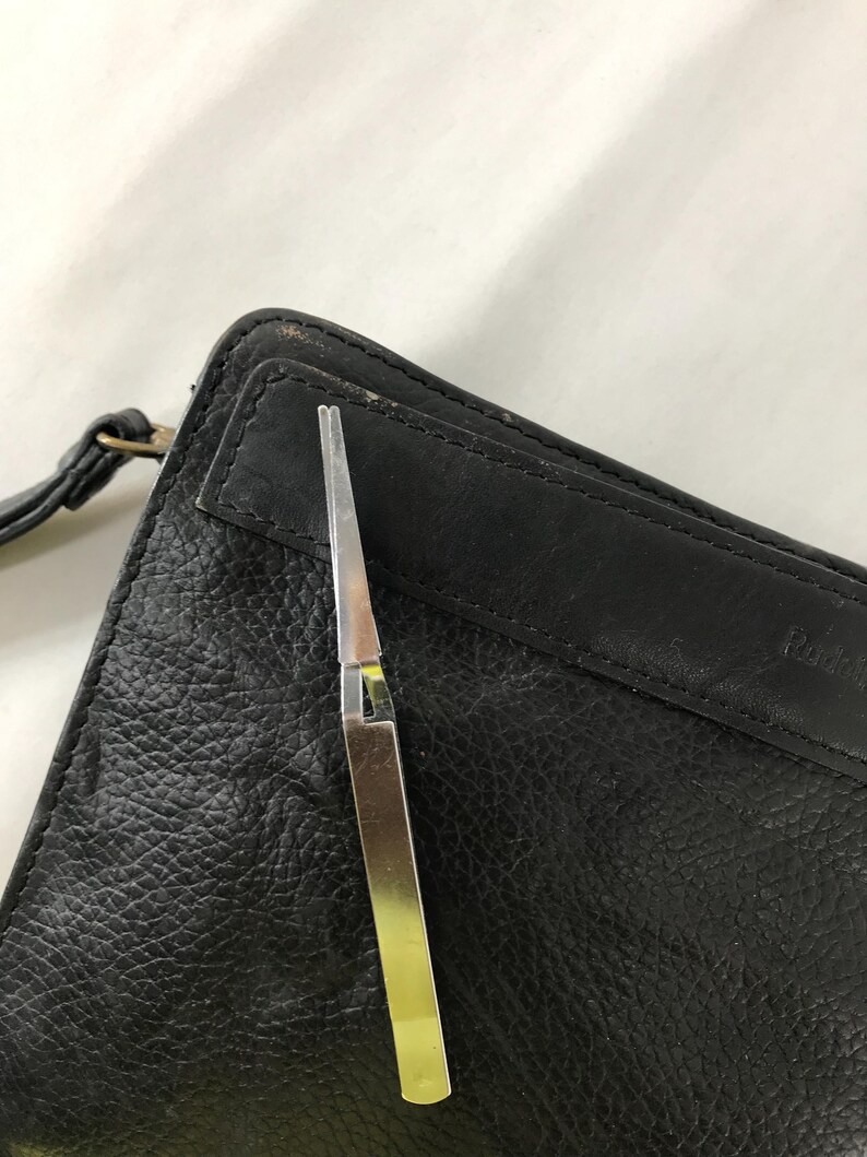 May include: A silver and gold metal tweezer tool is positioned over a black leather wallet. The wallet has a textured surface and a strap. The tweezer is open, revealing its pointed tips.