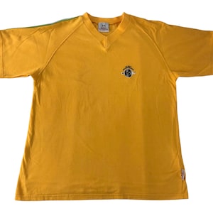 May include: Mustard yellow V-neck t-shirt with short sleeves. The shirt has a small emblem on the chest and a green stripe detail on the sleeves. The shirt is made of a soft material.