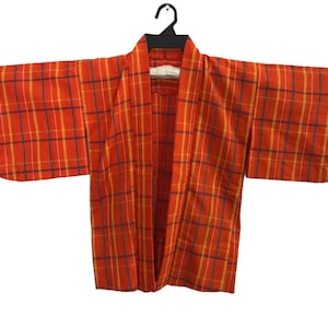 May include: An orange kimono with a plaid pattern in yellow and navy blue. The kimono is hanging on a black hanger and a red rod, against a white background. The sleeves are wide and the fabric appears lightweight.