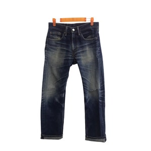Könnte beinhalten: Dunkelblaue Jeans mit einem aufgerissenen Knie Detail, hängen an einem Holzbügel. Die Jeans haben ein klassisches Fünf-Taschen-Design und einen Knopf- und Reißverschluss. Der Denim hat verblasste Bereiche.