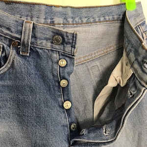 May include: A pair of light blue denim jeans with a button fly and five metal buttons. The jeans are unzipped and the waistband is folded over.