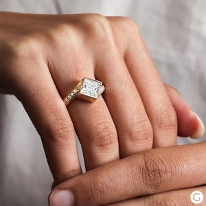 May include: A gold engagement ring with a large square-shaped diamond set in a unique, angled setting. The band is adorned with smaller diamonds.