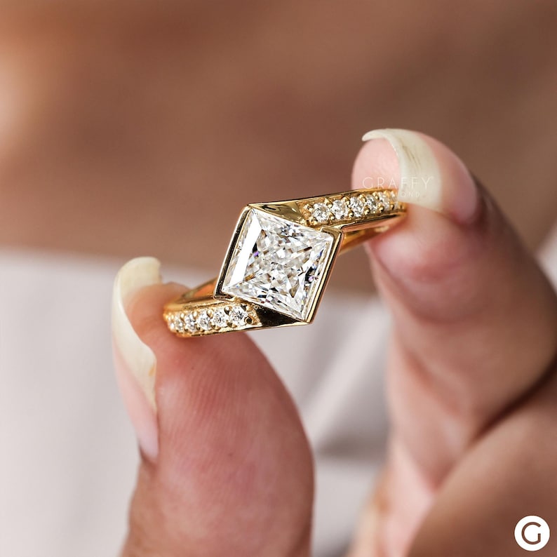 May include: A gold engagement ring with a square-shaped diamond set in a unique, angled setting. The band is adorned with smaller diamonds.