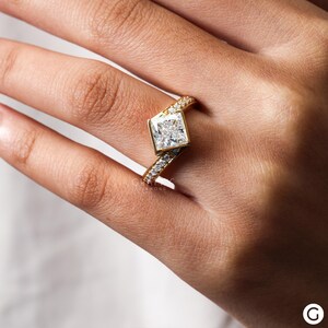 May include: A gold engagement ring with a large, square-shaped diamond set at an angle. The band is adorned with smaller diamonds.