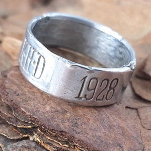 May include: A silver ring, engraved with the year "1928" and the word "UNITED". The ring is displayed on a textured, brown bark surface. The ring appears to be made of metal.