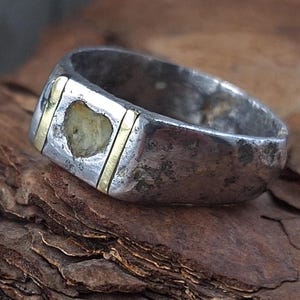 Può includere: Un anello d'argento con una faccia rettangolare, con una pietra a forma di cuore e dettagli dorati. L'anello ha un aspetto usurato e vintage, con segni di usura visibili. L'anello è appoggiato su una superficie di legno marrone strutturata.