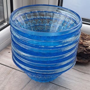 Op de afbeelding: Een blauwe glazen kom met horizontale strepen. De kom heeft een brede, open bovenkant en een licht taps toelopende basis. Het glas heeft een textuurpatroon aan de binnenkant. De kom staat op een houten oppervlak.