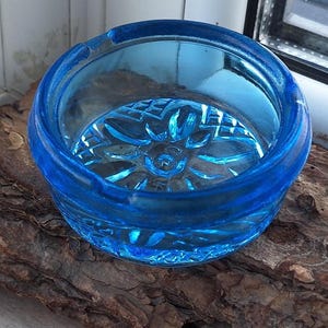 Op de afbeelding: Een ronde, helderblauwe glazen asbak met een bloemmotief op de bodem. De asbak staat op een stuk ruwe, bruine schors.