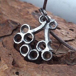 May include: A silver-colored pendant with a unique, openwork design featuring multiple circular openings. The pendant is suspended from a brown cord, creating a rustic and artistic aesthetic. The pendant's intricate details suggest it is handmade.