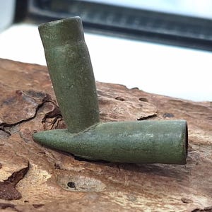 Pode incluir: Um cachimbo artesanal verde-oliva feito de um estojo de bala reaproveitado. O cachimbo apresenta uma tigela cilíndrica e uma haste angulada em aproximadamente 90 graus. O metal tem uma aparência desgastada.