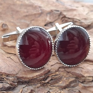 Pode incluir: Um par de abotoaduras prateadas com pedras cabochão ovais, vermelho escuro. As pedras são colocadas numa moldura ondulada. As abotoaduras são apresentadas numa superfície de madeira castanha texturizada.