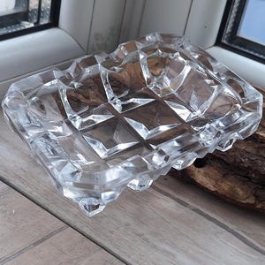 Könnte beinhalten: Klarer, rechteckiger Aschenbecher mit geometrischem, facettiertem Design. Der Aschenbecher besteht aus Glas und hat eine dekorative, strukturierte Oberfläche. Die Kanten sind leicht gewellt. Ein Stück Holz befindet sich neben dem Aschenbecher.