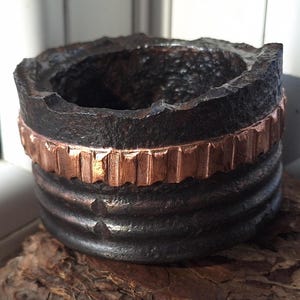 Puede incluir: Un recipiente cilíndrico, hecho a mano, de color marrón oscuro con una superficie texturizada y una banda de color cobre. El borde tiene un borde áspero e irregular. El recipiente está sobre un trozo de madera.