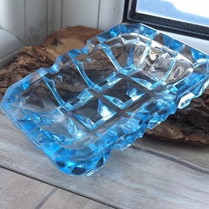 Könnte beinhalten: Ein quadratischer, hellblauer Glasaschenbecher mit einem geometrischen Rautenmuster. Der Aschenbecher besteht aus dickem Glas und hat eine strukturierte Oberfläche. Er liegt auf einem Stück Holz.