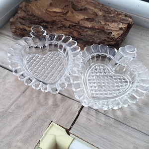May include: Two clear, heart-shaped glass dishes with a textured interior and scalloped edges. The dishes are displayed on a light-colored wooden surface, with a piece of bark in the background.