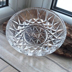 Puede incluir: Un cuenco de cristal transparente de forma circular con un detallado patrón geométrico tallado. El diseño incluye un motivo central de estrella rodeado de facetas repetitivas en forma de rombos y triángulos hasta el borde festoneado.