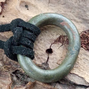 Op de afbeelding: Een groene metalen ring met een versleten oppervlak, bevestigd aan een zwart koord. De ring is met een knoop aan het koord vastgemaakt.