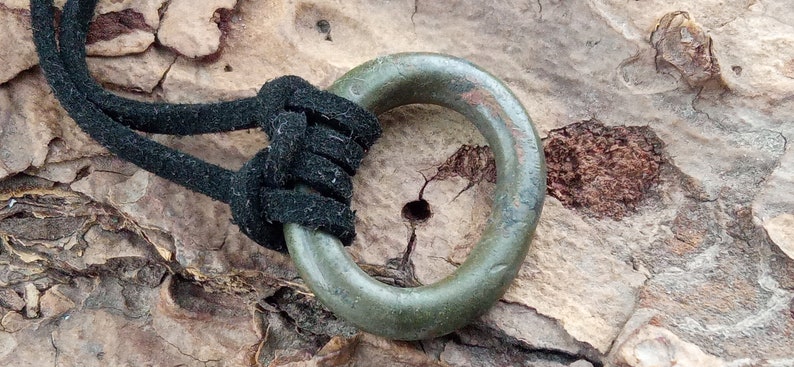 Op de afbeelding: Een groene metalen ring met een versleten oppervlak, bevestigd aan een zwart koord. De ring is met een knoop aan het koord vastgemaakt.
