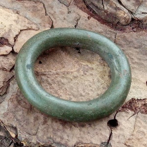 Op de afbeelding: Een groene metalen ring, mogelijk een oud artefact, liggend op een getextureerd bruin oppervlak.