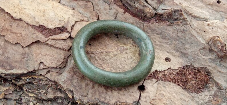 Op de afbeelding: Een groene metalen ring, mogelijk een oud artefact, liggend op een getextureerd bruin oppervlak.