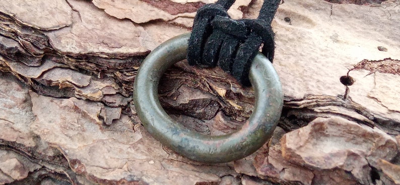 Op de afbeelding: Een grote, ronde, groene metalen ringhanger met een zwart koord eraan bevestigd. De hanger heeft een glad, versleten oppervlak en hangt aan een stuk boomschors.