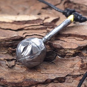 May include: A silver-colored pendant necklace featuring a textured, spherical charm with carved details. The charm is attached to a silver bar and a black cord. The necklace has a rustic, handcrafted appearance.