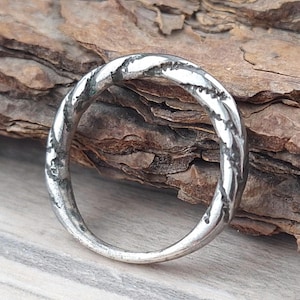 Puede incluir: Un anillo plateado con un diseño retorcido. El anillo tiene una superficie texturizada y está colocado sobre un fondo de madera y una superficie gris. El diseño del anillo es un bucle continuo.