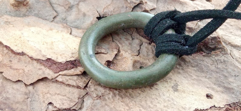 Op de afbeelding: Een groene metalen ring met een zwart koord erdoorheen geknoopt. De ring ligt op een ruw, bruin oppervlak.