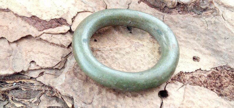 Op de afbeelding: Een groene metalen ring, mogelijk van brons, met een glad, afgerond oppervlak. De ring ligt op een stuk schors.
