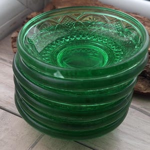 Op de afbeelding: Een stapel van zes groene glazen kommen. Elke kom heeft een cirkelvorm met een iets verhoogde rand en een decoratief patroon aan de binnenkant. De kommen zijn op elkaar gestapeld en tonen hun levendige groene kleur.